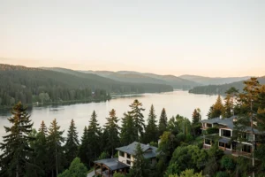 Elevated view of a broad calm river in the Pacific Northwest at golden hour, with upscale contemporary homes set among evergreens on a forested bluff